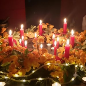 May include: A circular wreath adorned with vibrant orange and yellow flowers, interspersed with multiple tall red pillar candles and smaller white tea light candles. A string of warm white fairy lights is woven throughout the foliage.