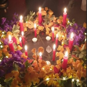 May include: A circular wreath decorated with purple and yellow flowers, green foliage, and small white string lights. Multiple red taper candles and two small white tea light candles are arranged around a central pentagram symbol and a clear crystal.
