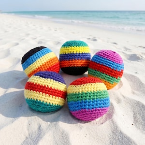 May include: Five brightly coloured, crocheted balls on a sandy beach. Each ball has a rainbow pattern of colours, including red, orange, yellow, green, blue, and purple. The balls are round and appear handmade.