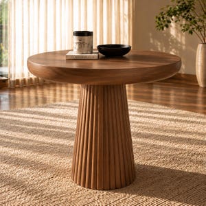 May include: A round wooden dining table with a thick, smooth top and a fluted, cylindrical pedestal base. On the table sits a white patterned cylindrical container resting on a book titled The Kinfolk and a small, plain black bowl.