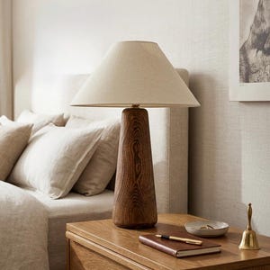 May include: A table lamp with a dark brown, tapered wooden base featuring a prominent wood grain pattern and a conical, off-white fabric lampshade. The lamp stands on a wooden surface alongside a small notebook, a pen, a dish, and a brass bell.