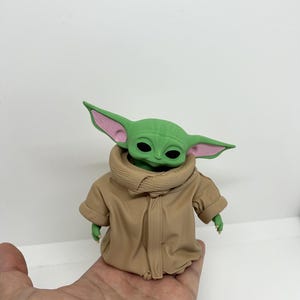 May include: A small green creature figurine with large pointed ears, black eyes, and a light pink inner ear detail. The figure is wearing a tan-coloured robe with a high, textured collar that wraps around the neck and covers the torso.