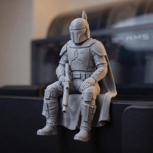Pode incluir: Uma estatueta cinzenta impressa em 3D de um guerreiro blindado sentado, usando um capacete com viseira em forma de T. A figura apresenta uma armadura de peito detalhada, um cinto utilit&aacute;rio, uma capa e segura uma espingarda no colo.