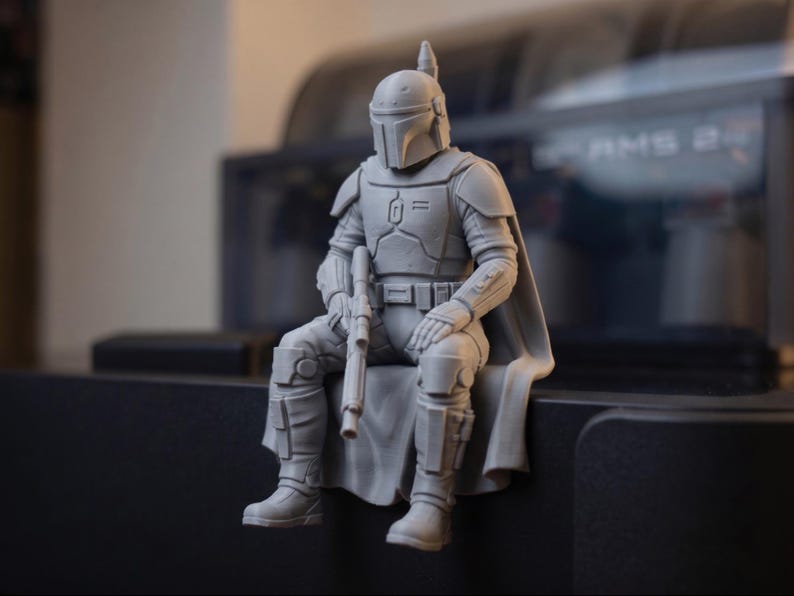 Pode incluir: Uma estatueta cinzenta impressa em 3D de um guerreiro blindado sentado, usando um capacete com viseira em forma de T. A figura apresenta uma armadura de peito detalhada, um cinto utilit&aacute;rio, uma capa e segura uma espingarda no colo.