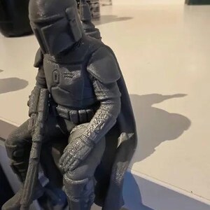 Pode incluir: Uma estatueta cinzenta impressa em 3D de um guerreiro com capacete, armadura de peito e capa, sentado numa posi&ccedil;&atilde;o sentada enquanto segura uma espingarda longa. A figura apresenta texturas detalhadas no fato, luvas e botas.
