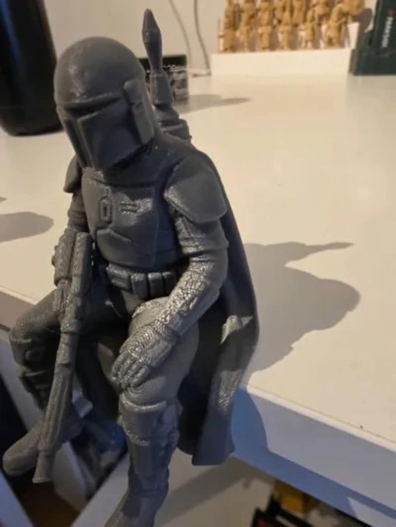 Pode incluir: Uma estatueta cinzenta impressa em 3D de um guerreiro com capacete, armadura de peito e capa, sentado numa posi&ccedil;&atilde;o sentada enquanto segura uma espingarda longa. A figura apresenta texturas detalhadas no fato, luvas e botas.