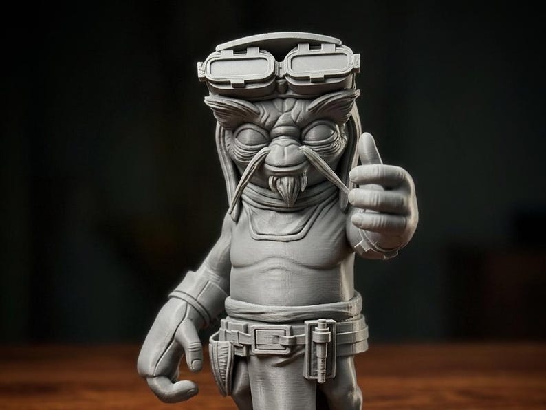 May include: A grey 3D printed figurine of a fictional creature with large ears, a goatee, and prominent eyebrows. The character wears goggles on its forehead, a belt with pouches, and is captured in a pose with one hand giving a thumbs-up gesture.