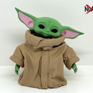 May include: A green creature figurine with large pointed ears featuring pink inner details and black eyes. The figure wears a tan hooded robe with a textured collar. The text NO AMS is visible in the top right corner with a red X over the O.