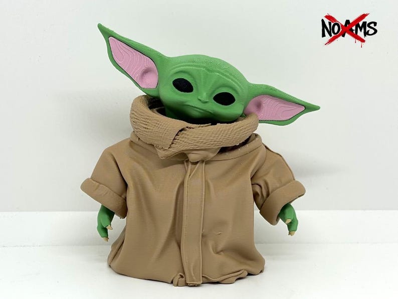 May include: A green creature figurine with large pointed ears featuring pink inner details and black eyes. The figure wears a tan hooded robe with a textured collar. The text NO AMS is visible in the top right corner with a red X over the O.