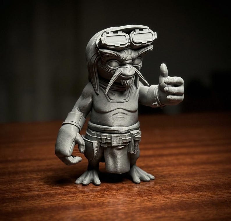 May include: A monochromatic grey figurine of a creature with large eyes, long whiskers, and a goatee. The figure wears goggles on its head, a belt with a pouch, and a loincloth. It is posed with one hand raised in a thumbs-up gesture.