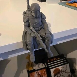 Pode incluir: Uma estatueta cinzenta impressa em 3D de um guerreiro com armadura e capacete, sentado com as pernas penduradas. A figura usa uma capa, um jetpack nas costas e segura uma espingarda longa no colo. A superf&iacute;cie mostra linhas de impress&atilde;o.