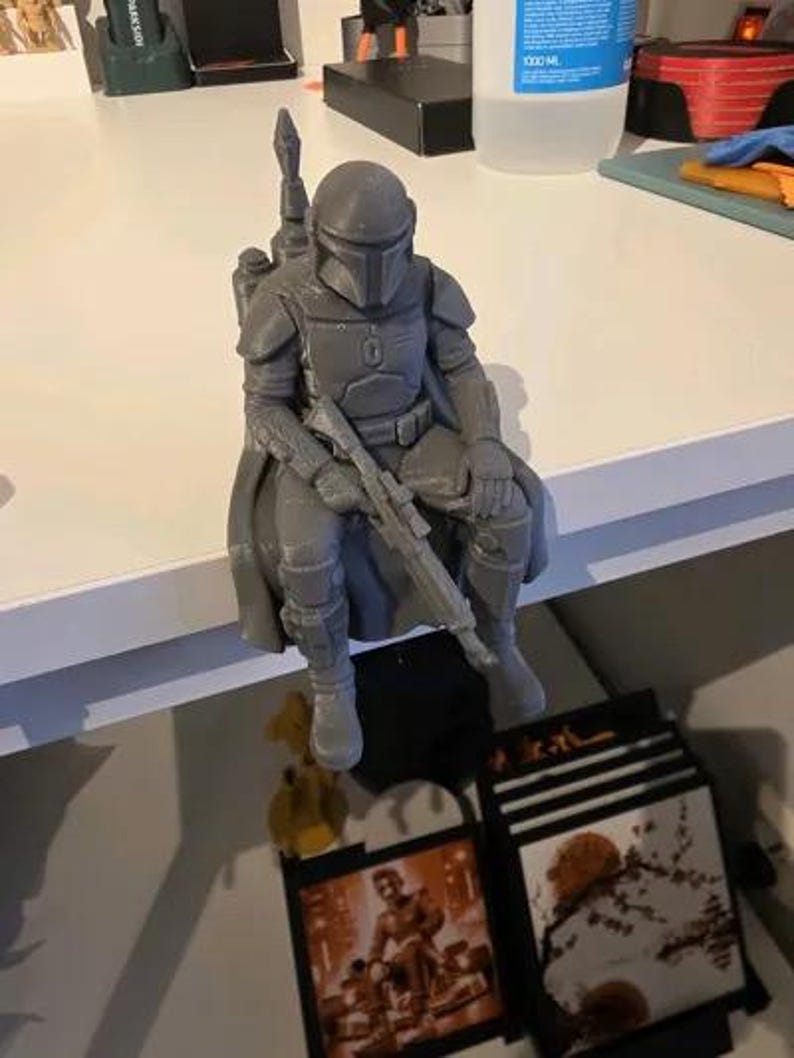 Pode incluir: Uma estatueta cinzenta impressa em 3D de um guerreiro com armadura e capacete, sentado com as pernas penduradas. A figura usa uma capa, um jetpack nas costas e segura uma espingarda longa no colo. A superf&iacute;cie mostra linhas de impress&atilde;o.
