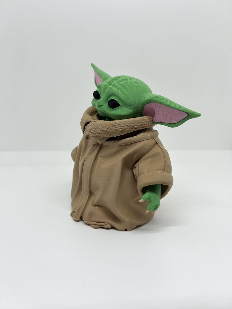 May include: A small green creature figurine with large pointed ears, black eyes, and pink inner ear details. The figure is wearing a tan-coloured robe with a textured collar and wide sleeves, showing small green hands with tiny claws extending from the garment.