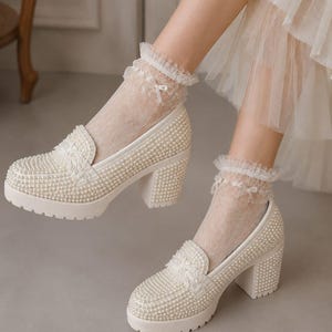 May include: A pair of cream-colored block-heeled loafers completely covered in small white faux pearls. The shoes feature a thick lug sole and a decorative lace-trimmed panel on the vamp. They are worn with sheer, lacy ankle socks with ruffled edges.