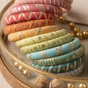 May include: A collection of colorful fabric-wrapped bangles in shades of pink, red, orange, yellow, green, blue, and teal. Each bangle is adorned with gold thread and sequins, arranged in a decorative pattern. The bangles are displayed in a round, wooden-rimmed tray.