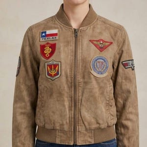 Könnte beinhalten: Eine hellbraune Bomberjacke mit geripptem Kragen und Saum sowie einem Frontreißverschluss. Die Jacke ist mit verschiedenen gestickten Aufnähern verziert, darunter eine Texas-Flagge mit dem Text TEXLAS, ein rotes Schild mit Schlange und ein Top Gun-Aufnäher.