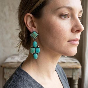 May include: Turquoise and silver earrings. The earrings feature a diamond-shaped top stone, followed by an oval stone, two square stones, and a final oval stone. The stones are a vibrant turquoise color, set in silver metal.