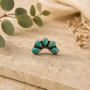 May include: A silver-toned ring featuring five turquoise teardrop-shaped stones. The stones are arranged in an arc, with a scalloped edge around each stone. The ring is set against a neutral background.