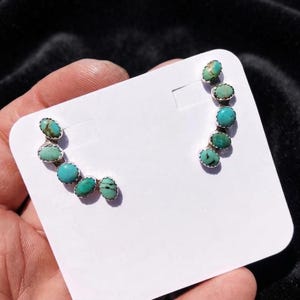 May include: A pair of ear climber earrings featuring five oval-shaped turquoise stones set in silver. Each earring is arranged in a curved line, showcasing natural blue-green variations and dark matrix patterns within the polished stone settings.