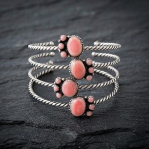 Puede incluir: Una pulsera tipo brazalete de múltiples hilos con cuatro bandas de plata trenzada. Cada banda está adornada con una piedra rosa ovalada central rodeada de piedras rosas redondas más pequeñas, creando un diseño de racimo en la parte frontal.
