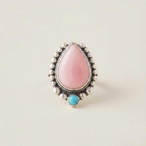 May include: A silver ring featuring a large, pear-shaped pink stone at the center, surrounded by a border of small silver spheres. A single, round, bright blue turquoise stone is set at the base of the pear-shaped stone, completing the design.