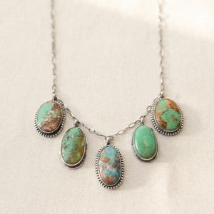 May include: A silver chain necklace featuring five oval-shaped turquoise stones set in silver bezels with beaded edges. The stones display natural variations of blue, green, and brown matrix patterns, suspended symmetrically along the delicate metal chain.
