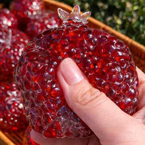 Puede incluir: Una granada de cristal rojo transparente con una superficie texturizada, sostenida en una mano. La granada tiene una parte superior transparente en forma de corona. Otras granadas son visibles en el fondo, en una cesta tejida.