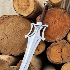 May include: A silver-coloured metal sword featuring a double-edged blade with a pointed tip and a stylised crossguard. The handle is wrapped in brown leather material, and the pommel is a simple silver-coloured metal cap at the end of the grip.