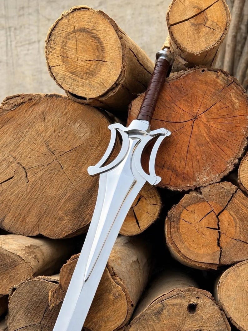 May include: A silver-coloured metal sword featuring a double-edged blade with a pointed tip and a stylised crossguard. The handle is wrapped in brown leather material, and the pommel is a simple silver-coloured metal cap at the end of the grip.