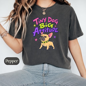 Könnte beinhalten: Ein anthrazitfarbenes Kurzarm-T-Shirt mit einer Grafik einer beigen französischen Bulldogge, die ein neongrünes Stachelhalsband trägt. Über dem Hund steht der Text Tiny Dog Big Attitude in pinken, neongrünen und violetten Buchstaben mit Sternen.