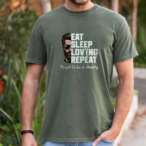 Peut inclure: Un t-shirt à manches courtes vert olive avec un motif représentant une personne barbu portant des lunettes de soleil. Le texte indique EAT SLEEP LOVING REPEAT et Proud to be a Daddy. Le motif est imprimé en blanc et noir sur le devant du t-shirt.