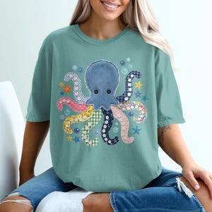 Puede incluir: Una camiseta de manga corta color verde salvia con un gráfico central de un pulpo con tentáculos estilo patchwork en varios patrones, incluyendo rayas, lunares y cuadros, rodeado de pequeñas burbujas azules y estrellas de mar naranjas y amarillas.