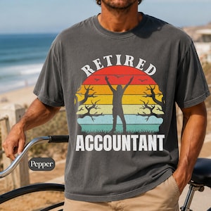 Può includere: Una t-shirt a maniche corte grigio antracite con una stampa grafica che riporta il testo "RETIRED ACCOUNTANT" in bianco. Il design include la sagoma di una persona con le braccia alzate su uno sfondo al tramonto a strisce rosse, arancioni, gialle e verde acqua.