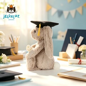 Puede incluir: Un peluche de conejo gris claro con un birrete negro con borla amarilla, sosteniendo un pergamino enrollado. El conejo est&aacute; sentado sobre una superficie blanca, con otros birretes y diplomas en el fondo. El logotipo de Jellycat London es visible.