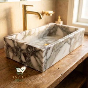 May include: Rectangular marble sink with a gold faucet. The sink has a white and gray veined pattern. The faucet and soap dish are gold. The sink is on a wooden surface. The text "TARIFF FREE" is visible.