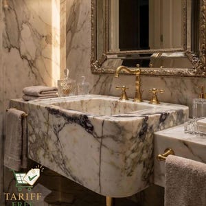 May include: A luxurious marble bathroom sink with gold fixtures. The sink has a rectangular basin and is set against a marble wall. A gold-framed mirror hangs above the sink, and towels are neatly arranged nearby.