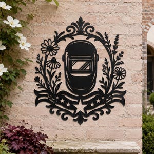 May include: A black metal wall art piece featuring a central welding helmet surrounded by a decorative frame of flowers, leaves, and a banner with stars at the bottom. The design is symmetrical with intricate scrollwork and floral elements.
