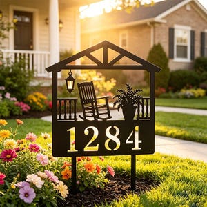 May include: A black metal house number sign featuring the digits 1284. The design includes a cutout silhouette of a porch with a rocking chair, a hanging lantern, and a potted plant, all framed within a house-shaped structure on two stakes.