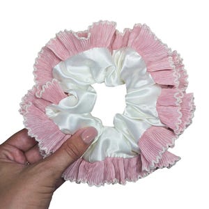 May include: A hair scrunchie featuring a smooth, off-white satin inner band surrounded by a pleated, light pink fabric ruffle. The outer edge of the pink ruffle is finished with a delicate, scalloped white trim, creating a layered, circular design.