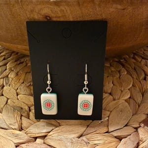 May include: A pair of drop earrings featuring rectangular white tiles with a green and red floral-like symbol, suspended from silver-toned metal hooks. The earrings are displayed on a black rectangular card with a small hole at the top.