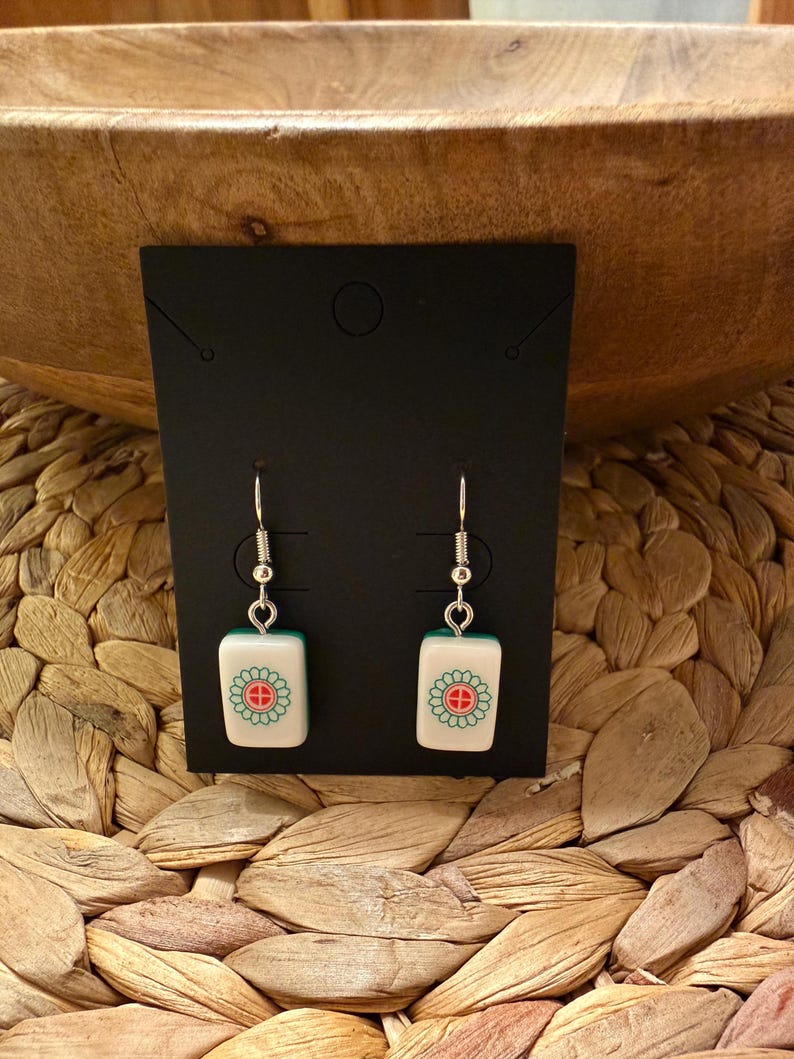 May include: A pair of drop earrings featuring rectangular white tiles with a green and red floral-like symbol, suspended from silver-toned metal hooks. The earrings are displayed on a black rectangular card with a small hole at the top.