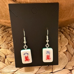May include: A pair of dangle earrings featuring white rectangular mahjong tiles as charms. Each tile displays blue and red Chinese characters. The charms are suspended from silver-toned metal fishhook wires and are attached to a black display card.