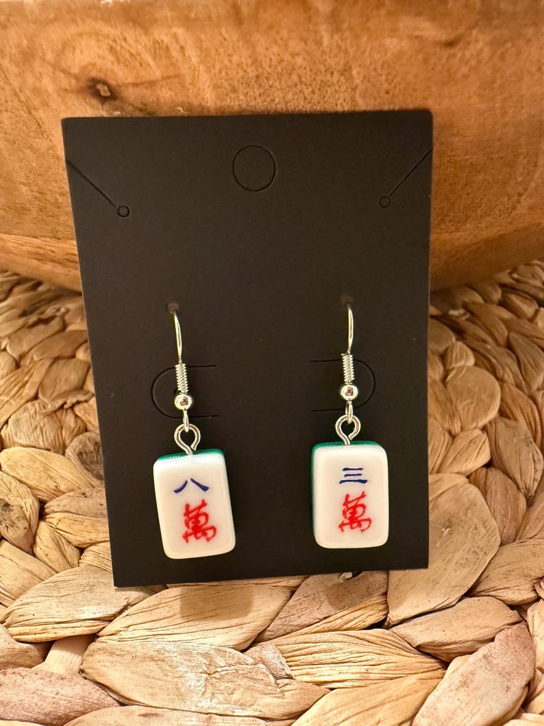 May include: A pair of dangle earrings featuring white rectangular mahjong tiles as charms. Each tile displays blue and red Chinese characters. The charms are suspended from silver-toned metal fishhook wires and are attached to a black display card.