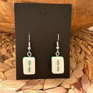 May include: A pair of dangle earrings featuring white rectangular mahjong tiles with a green two-bamboo symbol. The tiles are attached to silver-toned metal fishhook ear wires, presented on a black rectangular display card.