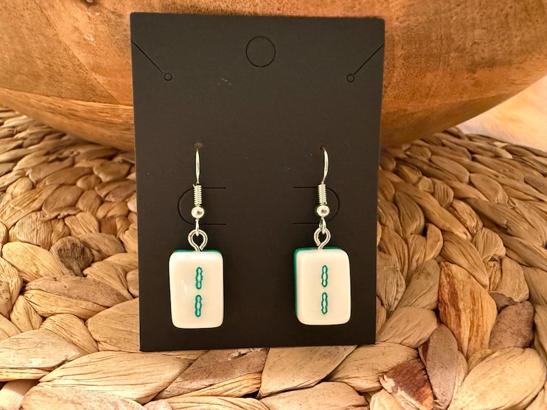 May include: A pair of dangle earrings featuring white rectangular mahjong tiles with a green two-bamboo symbol. The tiles are attached to silver-toned metal fishhook ear wires, presented on a black rectangular display card.