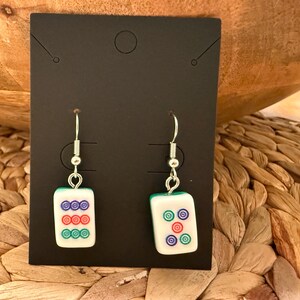 May include: A pair of dangle earrings featuring rectangular white mahjong tiles with green backs. The left earring displays three rows of three colored circles in purple, red, and green. The right earring shows five colored circles in a cross pattern.