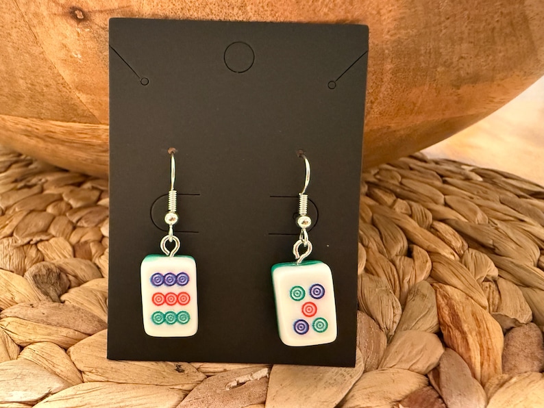 May include: A pair of dangle earrings featuring rectangular white mahjong tiles with green backs. The left earring displays three rows of three colored circles in purple, red, and green. The right earring shows five colored circles in a cross pattern.