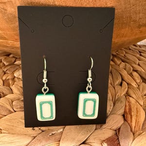 May include: A pair of dangle earrings featuring white and green rectangular mahjong tiles as charms. The tiles are suspended from silver-toned metal fishhook wires and are displayed on a rectangular black card with a top center hole.