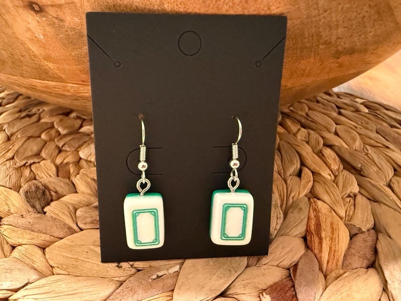 May include: A pair of dangle earrings featuring white and green rectangular mahjong tiles as charms. The tiles are suspended from silver-toned metal fishhook wires and are displayed on a rectangular black card with a top center hole.