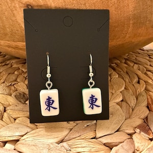 May include: A pair of dangle earrings featuring white mahjong tiles with the blue Chinese character for East, suspended from silver-toned metal hooks. The earrings are displayed on a rectangular black card with a circular hole at the top.